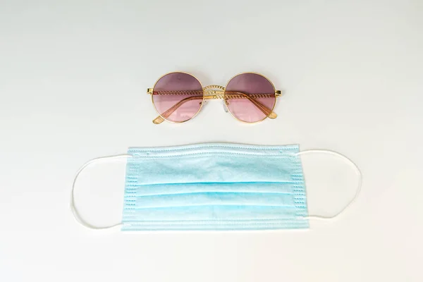 Sunglasses and a blue face medicine mask on a white background. Flat lay composition with summer vacation accessories. Travel trends and rules in 2020 due to coronavirus