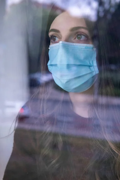 Young caucasian woman with nostalgic face looking out the window and wearing a face mask. Covid 19. Social distance.