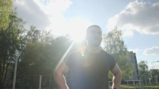 Close up Forte fisiculturista muscular com barba caminha ao redor do estádio contra a luz de fundo e céu azul . — Vídeo de Stock