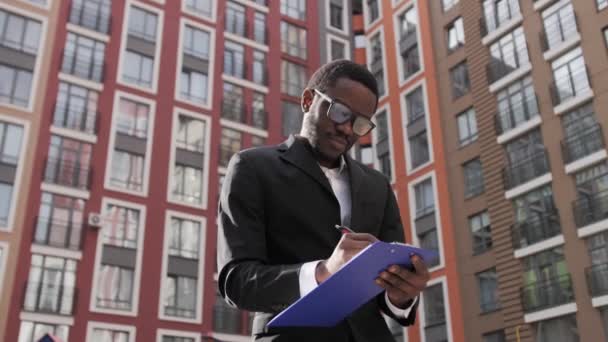 Cute black engineer with glasses holding a blue tablet for paper on a background of beautiful houses. — Stock Video