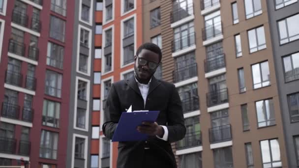 Αφρο-Αμερικανός μηχανικός με γυαλιά κρατώντας ένα μπλε δισκίο για το χαρτί σε ένα φόντο των όμορφων σπιτιών. — Αρχείο Βίντεο