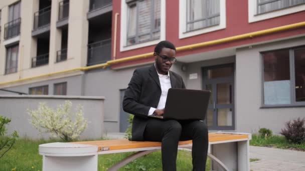 Schöner schwarzer Geschäftsmann im schwarzen Anzug, der draußen auf einer Bank sitzt und an einem Laptop arbeitet. — Stockvideo