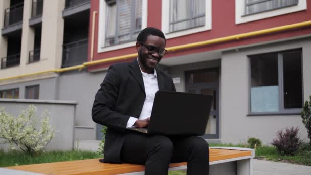黒人のスーツを着たアフリカ系アメリカ人ビジネスマンが屋外のベンチに座ってノートパソコンで仕事をしている. — ストック動画