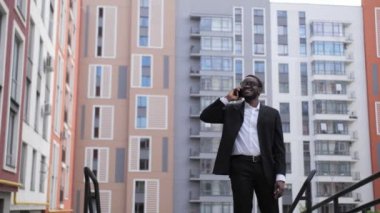 Siyah takım elbiseli ve gözlüklü Afro-Amerikan işadamı dışarıda telefonla konuşuyor, güzel renkli evlerin arka planında gülümsüyor..