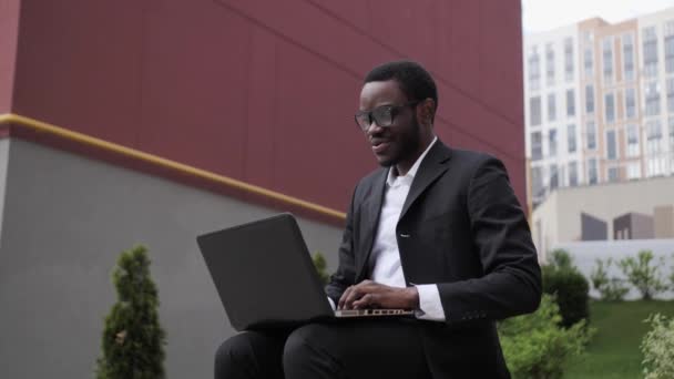 屋外のベンチに座っている黒いスーツを着たハンサムな黒人ビジネスマンは、ビデオ通話やラップトップでの作業にWebカメラを使用します。. — ストック動画