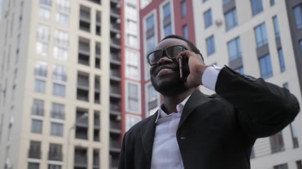 Handsome black businessman in a black suit and glasses talking on the phone outdoors on the background of beautiful colored houses.Close up. — Stock Video
