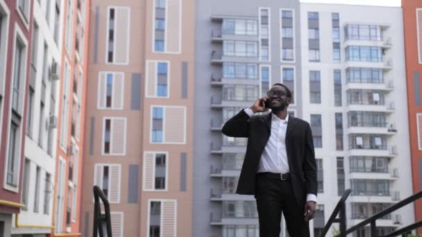 Αφρο Αμερικανός επιχειρηματίας με μαύρο κοστούμι και γυαλιά μιλώντας στο τηλέφωνο σε εξωτερικούς χώρους, χαμογελώντας στο παρασκήνιο των όμορφων χρωματιστά σπίτια. — Αρχείο Βίντεο