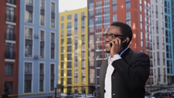 Afro American manager in a black suit and glasses talking on the phone outdoors on the background of beautiful colored houses. — Stock Video