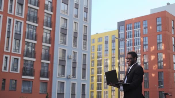 Όμορφος μαύρος επιχειρηματίας με μαύρο κοστούμι και γυαλιά που εργάζεται σε ένα φορητό υπολογιστή, χαμογελώντας, χρησιμοποιεί βιντεοκλήση σε ένα φορητό υπολογιστή στο παρασκήνιο των όμορφων νέων κτιρίων. — Αρχείο Βίντεο