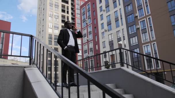 Afro American businessman in a black suit and glasses talking on the phone outdoors,smiling on the background of beautiful colored houses. — Stock Video