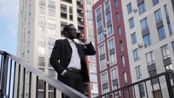 Donkere zakenman in een zwart pak en een bril die buiten aan de telefoon praat, glimlachend op de achtergrond van prachtige gekleurde huizen. — Stockvideo