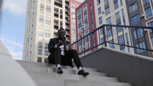 Przystojny afro Amerykanin siedzi na schodach z kawą i rozmawia na wideo link. — Wideo stockowe