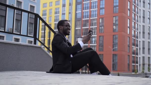 Vista da vicino di Bello un afro americano dalla pelle scura seduto sulle scale con caffè e parlando sul collegamento video . — Video Stock