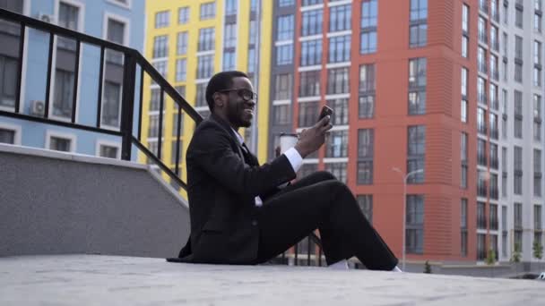 Vista de cerca de Guapo un afroamericano de piel oscura sentado en las escaleras con café y hablando en enlace de vídeo . — Vídeo de stock