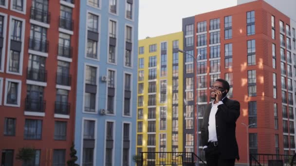 Hombre de negocios afroamericano guapo en un traje negro y gafas hablando por teléfono al aire libre, sonriendo en el fondo de hermosas casas de colores . — Vídeos de Stock