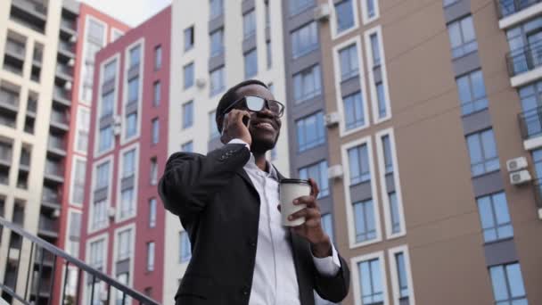 Snygg svart afro amerikan man talar i telefon och ler medan du dricker kaffe från en vit kopp på bakgrunden av vackra hus. — Stockvideo