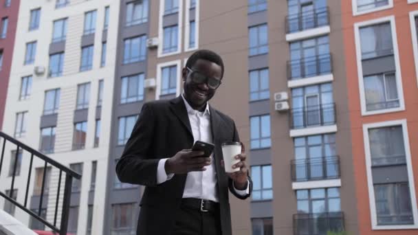 Netter afroamerikanischer Mann schreibt Nachricht am Telefon und lächelt, während er Kaffee aus einer weißen Tasse vor dem Hintergrund schöner Häuser trinkt. — Stockvideo