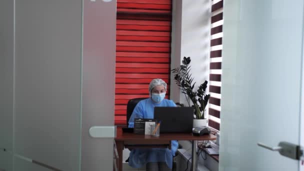 Γιατρός κάθεται σε ένα τραπέζι με ιατρικά ρούχα και μάσκα κατά τη διάρκεια μιας πανδημίας κορωναϊού σε μια κλινική ομορφιάς. — Αρχείο Βίντεο