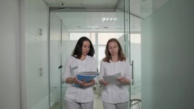 İki doktor klinikte ellerinde belgelerle yürür ve iş meselelerini tartışırlar..