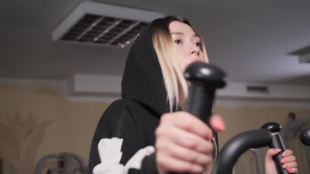 Pretty girl in a black jacket with a hood runs on the orbitrek in the gym.Woman trains in gym. Physical activity helps burn up calories. The woman goes in for sports. — Stock Video