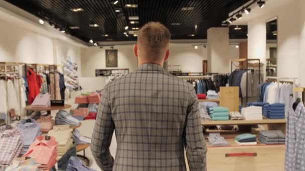 Empresario en un traje gris entra en la vista de la tienda de ropa de los hombres desde la parte posterior en el fondo de la tienda . — Vídeos de Stock