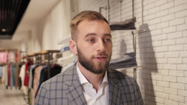 Retrato de un hombre de negocios con barba en una tienda de ropa masculina, de pie con ropa sobre fondos . — Vídeos de Stock