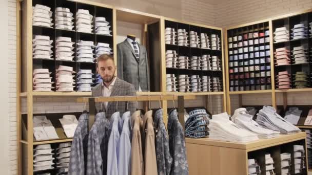 Boutique de ropa masculina de marca, hombre de negocios en un traje con barba se acerca al estante con camisas . — Vídeos de Stock