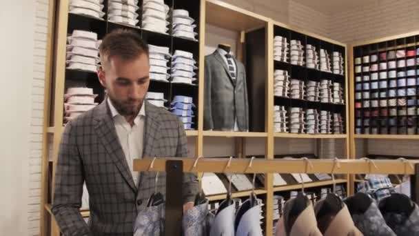 Boutique της μάρκας ανδρικά ρούχα, κομψό άνδρα σε ένα κοστούμι με γενειάδα πλησιάζει το ράφι με πουκάμισα. — Αρχείο Βίντεο