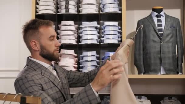 Boutique της μάρκας ανδρικά ρούχα, Κομψό άνθρωπος σε ένα γκρι κοστούμι με γενειάδα επιλέγει ένα πουκάμισο. — Αρχείο Βίντεο