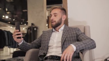 Gri takım elbiseli ve sakallı şık bir işadamı erkek giyim mağazasında akıllı telefon kullanarak görüntülü görüşme yapıyor..