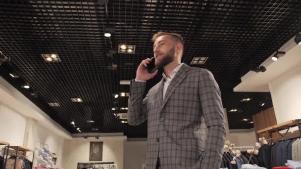 Elegante hombre de negocios en un traje gris y barba hablando por teléfono en una tienda de ropa para hombres . — Vídeos de Stock