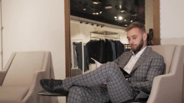 Gri takım elbiseli ve sakallı şık bir adam bir sandalyede oturuyor ve bir erkek giyim mağazasında günlük arıyor..