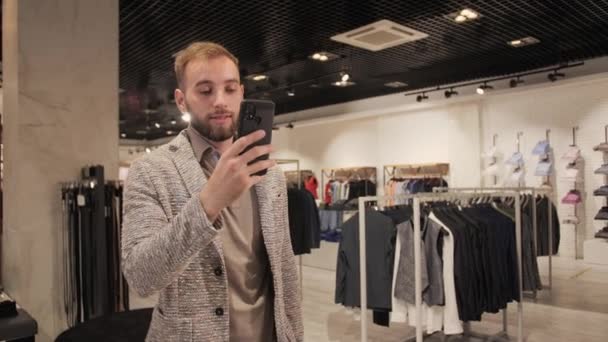 Stijlvolle knappe zakenman in pak en baard in gesprek op video met behulp van smartphone in een herenkledingwinkel. — Stockvideo