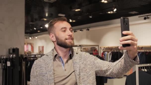 Elegante hombre de negocios guapo en un traje y barba hablando en videollamada usando un teléfono inteligente en una tienda de ropa para hombres . — Vídeos de Stock