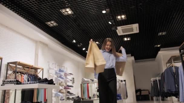 Menina feliz com pacotes em suas mãos danças em uma elegante loja de roupas de marca depois de fazer compras . — Vídeo de Stock