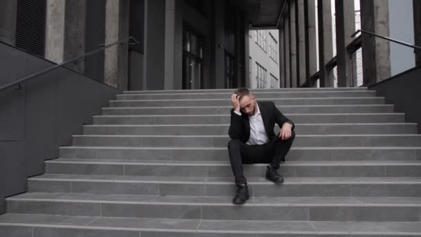 An fired office worker sits on the stairs and holds his head in crisis. — Stock Video