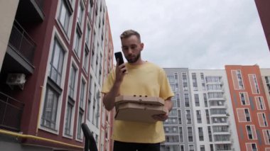 Sarı tişörtlü kurye çevrimiçi pizza teslimatı yapıyor. Servis elemanı, müşteriyi renkli evlerin arka planından arıyor..