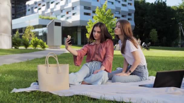 Zwei schöne Freundinnen sitzen draußen auf dem Gras und machen Selfies vor dem Hintergrund der Universität. Sie haben Spaß. — Stockvideo