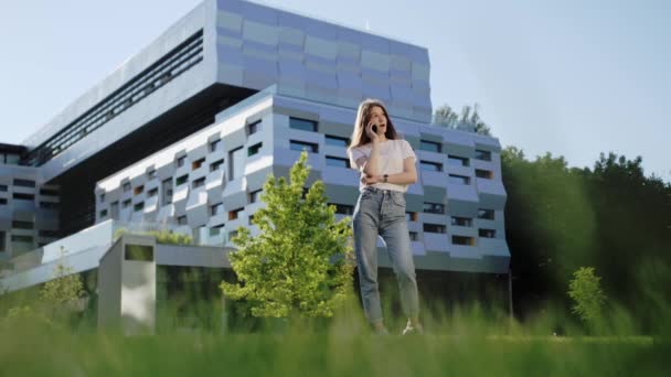 Mooi meisje in een wit t-shirt en jeans praten aan de telefoon met een vriend op de achtergrond van een modern gebouw. — Stockvideo