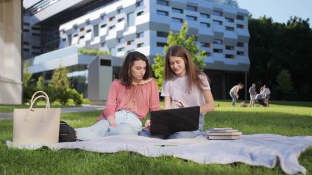 Duas estudantes bonitas estão estudando uma palestra remotamente em um laptop ao ar livre no fundo da universidade . — Vídeo de Stock