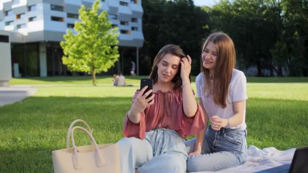 Deux belles copines assises sur l'herbe et parlant un appel vidéo elles saluent en saluant en souriant. — Video