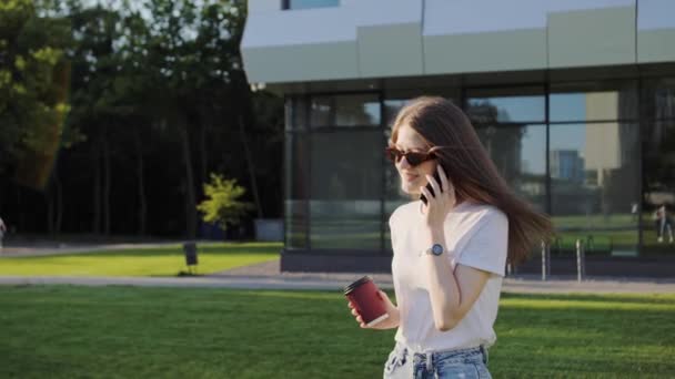 Mooi meisje in wit T-shirt met koffie aan de telefoon met een vriend op de achtergrond van een modern gebouw. Zonnige dag zomer. — Stockvideo