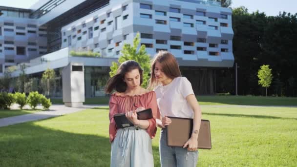 Duas namoradas femininas ficam perto da universidade com um laptop e um notebook e resolvem os trabalhos de casa remotamente . — Vídeo de Stock