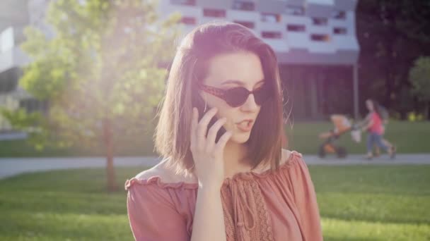 Modern bir binanın arka planında gülümseyen bir arkadaşıyla telefonda konuşan güzel bir kız. Güneşli yaz. — Stok video