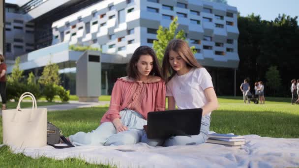 Två vackra kvinnliga studenter studerar en föreläsning på distans på en bärbar dator utomhus i bakgrunden av universitetet. — Stockvideo