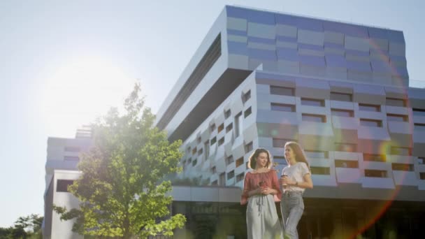 Twee mooie vrouwelijke studenten staan naast de universiteit met een laptop en een notitieboekje en lossen huiswerk op afstand op. — Stockvideo