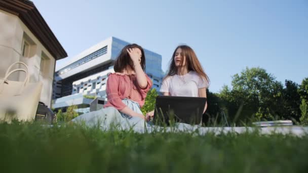 Duas estudantes bonitas estão estudando uma palestra remotamente em um laptop ao ar livre no fundo da universidade . — Vídeo de Stock