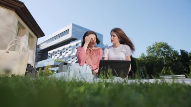 Dos bellas estudiantes femeninas están estudiando una conferencia remotamente en un portátil al aire libre en el fondo de la universidad . — Vídeos de Stock