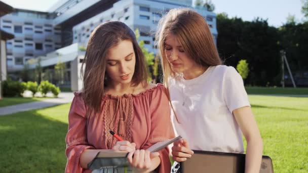 Zwei hübsche Freundinnen vom College bei Hausaufgaben im Freien vor dem Hintergrund eines modernen Gebäudes. — Stockvideo