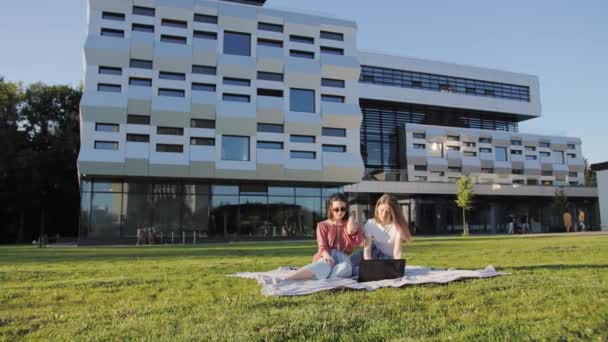 Duas estudantes atraentes do sexo feminino estão estudando uma palestra remotamente em um laptop ao ar livre no fundo do edifício moderno . — Vídeo de Stock
